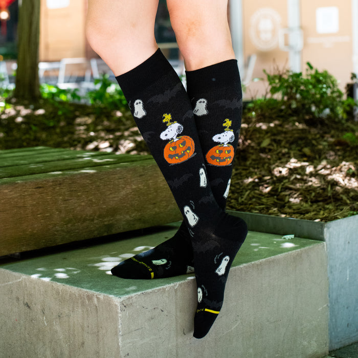 Black knee-high compression socks with a Peanuts mountain hike design, worn in an outdoor setting.